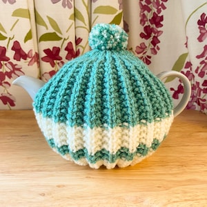 May include: Teal and white knitted teapot cosy with a white pom pom on top.