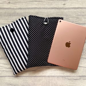 Op de afbeelding: Een iPad ligt naast twee tablethoezen. Eén hoes heeft zwart-witte verticale strepen, de andere heeft een zwarte achtergrond met witte stippen. De iPad is roségoud van kleur.