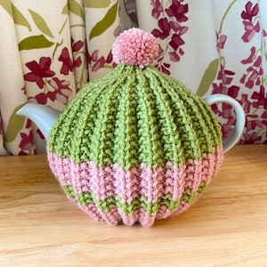 May include: A green and pink knitted tea cosy with a pom-pom on top, covering a white teapot. The cosy has a ribbed knit design. Floral curtains are visible in the background. The tea cosy is a handmade item.