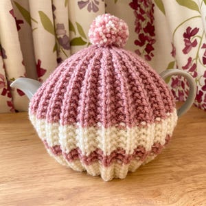 Op de afbeelding: Een roze en witte gebreide theemuts met een pompon erop. De theemuts bedekt een grijze theepot en is gemaakt van geribbelde gebreide stof. De theemuts staat op een houten oppervlak.