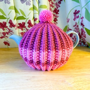 Puede incluir: Un cubretejera de punto en tonos rosas y morados, con un diseño acanalado y un pompón en la parte superior. Cubre una tetera azul claro. El fondo presenta cortinas florales.