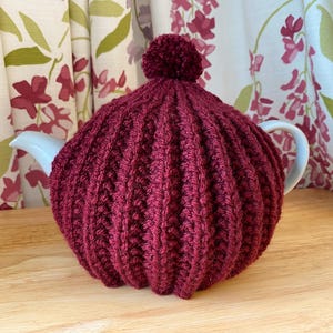 Puede incluir: Un cubreteiera de punto burdeos con diseño acanalado y un pompón en la parte superior. Cubre una tetera blanca con asa y boquilla curvadas. El fondo presenta cortinas florales y una superficie de madera.