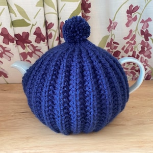 May include: A navy blue knitted tea cozy with a ribbed design and a pom-pom on top. The cozy is covering a white teapot with a curved handle. The background features floral curtains and a wooden surface.