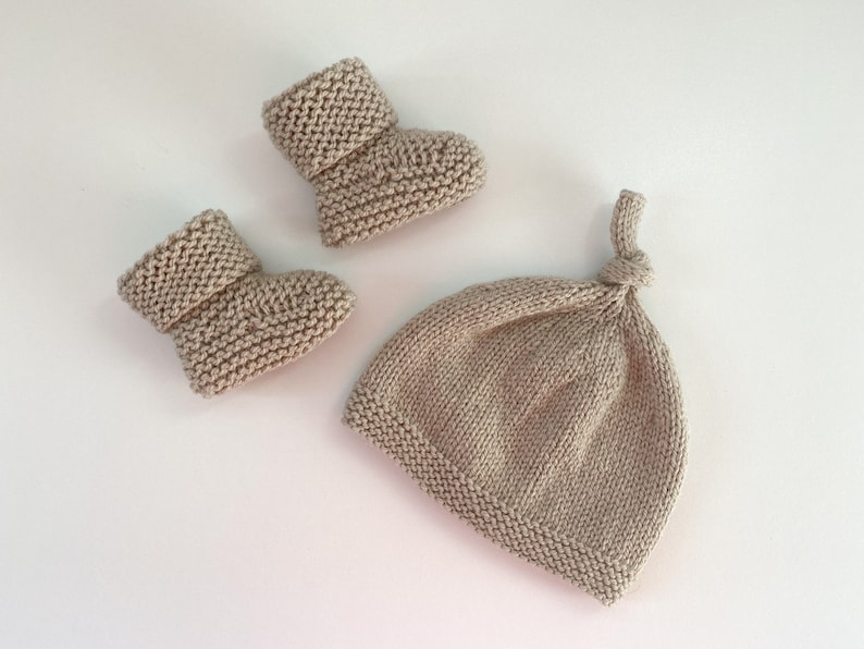 May include: A set of knitted baby booties and a matching hat in a light beige colour. The booties have a folded cuff and the hat has a knotted top.