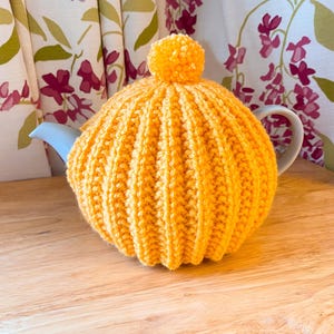 Può includere: Un scaldino per tè giallo lavorato a maglia con un pompon in cima, che copre una teiera azzurra. Lo scaldino ha un design a coste e poggia su una superficie di legno. Tende a motivi floreali sullo sfondo.