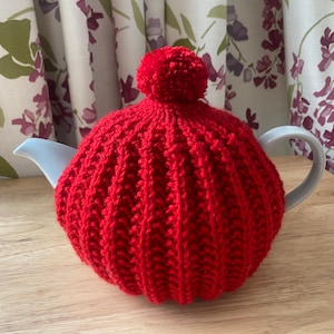 Könnte beinhalten: Ein roter Strick-Teewärmer mit einem Bommel oben, der eine weiße Teekanne bedeckt. Der Teewärmer hat ein Rippenmuster. Die Teekanne steht auf einer Holzoberfläche, mit floralen Vorhängen im Hintergrund.