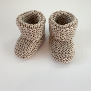 May include: A pair of beige knitted baby booties. The booties are made with a simple stitch pattern and have a fold-over cuff.