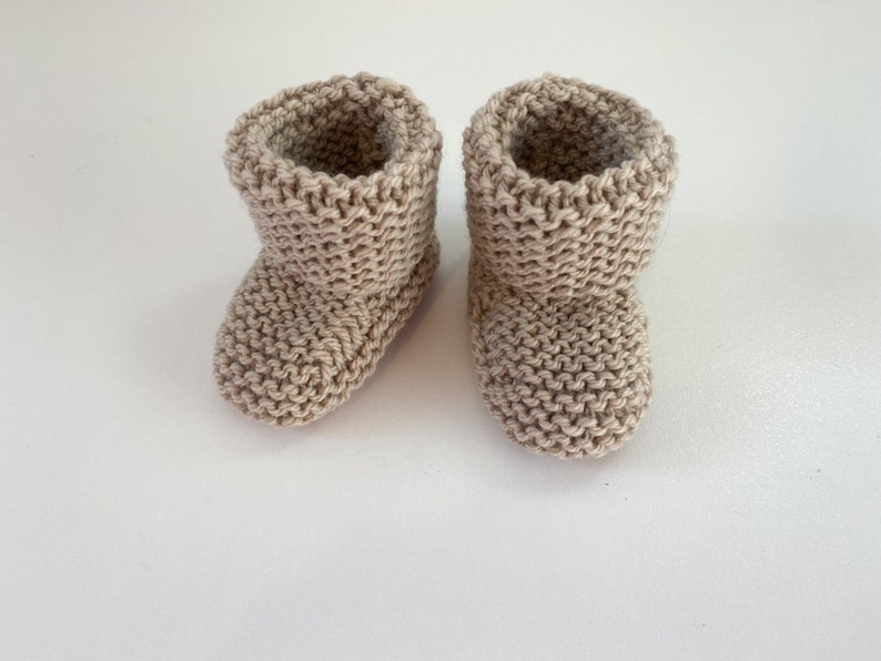May include: A pair of beige knitted baby booties. The booties are made with a simple stitch pattern and have a fold-over cuff.