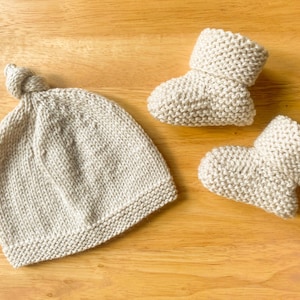May include: A light beige knitted baby hat with a knotted top and two matching baby booties. The set is made of a soft, textured yarn and is displayed on a wooden surface. The booties have folded-over cuffs.