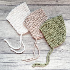 Può includere: Tre cappellini per neonati lavorati a maglia, in colori tenui: bianco sporco, rosa chiaro e verde salvia. Ogni cappellino ha un motivo a punto strutturato e lacci con filo coordinato. I cappellini sono esposti su una superficie di legno.