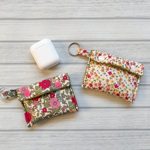 Op de afbeelding: Twee AirPod-hoesjes met bloemenprint met sleutelringen en een witte AirPod-oplaadcase. De hoesjes hebben een kleurrijk bloemenpatroon op een crèmekleurige achtergrond, met roze, rode en gele bloemen. De hoesjes worden gesloten met een drukknop.