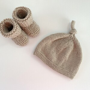 May include: A set of knitted baby booties and a matching hat in a light beige colour. The hat has a knotted top.