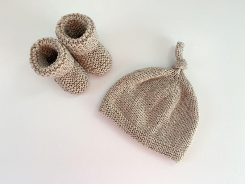 May include: A set of knitted baby booties and a matching hat in a light beige colour. The hat has a knotted top.