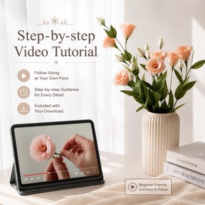 May include: A tablet displaying a step-by-step video tutorial on crafting a pink flower, positioned next to a ribbed cream vase containing a bouquet of artificial peach-coloured flowers and two stacked books titled The Kinfolk and Live Beautifully.