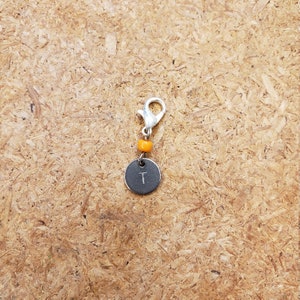 Puede incluir: Un pequeño dije plateado con un cierre de mosquetón y un colgante redondo gris oscuro. El colgante presenta la letra "T" y está conectado al cierre por una cuenta naranja. El dije está diseñado para un llavero o pulsera.