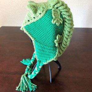 May include: Green crocheted hat with a dinosaur design. The hat has a long tail with a braided design and two tassels.