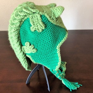 May include: Green crocheted hat with a three-dimensional design resembling a frog. The hat has a long tassel and is displayed on a black stand.