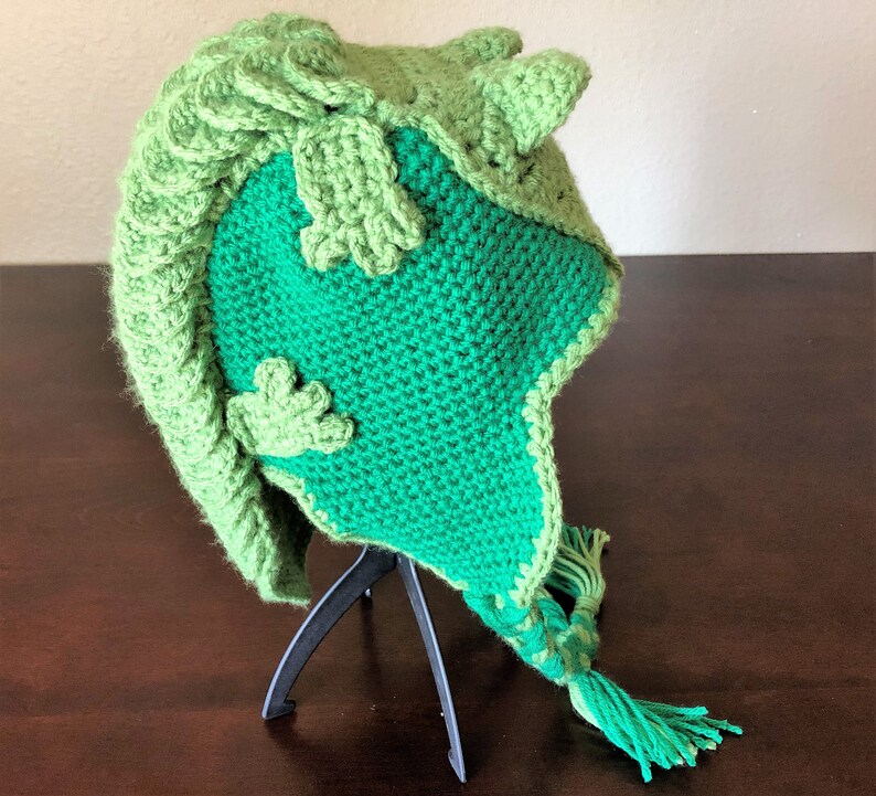 May include: Green crocheted hat with a three-dimensional design resembling a frog. The hat has a long tassel and is displayed on a black stand.