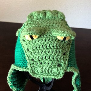 May include: Green crocheted alligator hat with yellow eyes and a green and dark green striped brim.