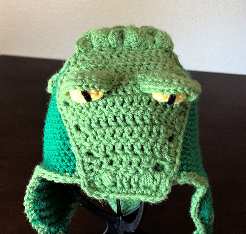 May include: Green crocheted alligator hat with yellow eyes and a green and dark green striped brim.