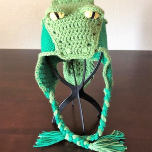 May include: Green crocheted alligator hat with long green and light green braided ties. The hat is displayed on a black hat stand.