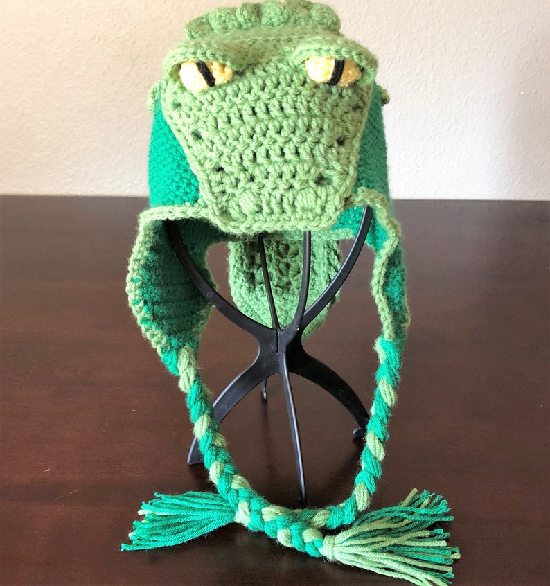 May include: Green crocheted alligator hat with long green and light green braided ties. The hat is displayed on a black hat stand.