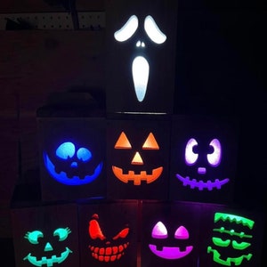 Pode incluir: Oito caixas de madeira com diferentes rostos temáticos de Halloween esculpidos nelas. As caixas são iluminadas por dentro com luzes de diferentes cores, incluindo azul, laranja, roxo, verde e branco.