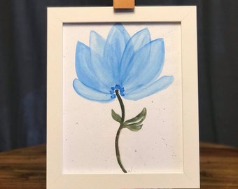 May include: A framed watercolor painting of a blue flower. The flower has delicate, translucent petals and a dark green stem with leaves. The artwork is displayed on a small wooden easel, with a white frame.
