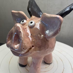 Può includere: Una scultura di maiale in ceramica rosa e nera. Il maiale ha grandi orecchie, un grugno e piccoli occhi rotondi. La scultura fatta a mano ha una finitura lucida. Il maiale è in piedi su quattro zampe corte.