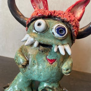 Könnte beinhalten: Eine Keramik-Monsterfigur mit einem texturierten grünen Körper, zwei großen Hörnern und einer roten Mütze. Sie hat ungleiche Augen, weiße Fangzähne und ein kleines rotes Herzdetail auf der Brust. Die Kreatur hat zwei Arme und zwei Füße mit dunklen Krallen.