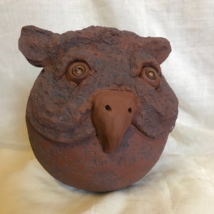 Könnte beinhalten: Eine Keramikskulptur eines Tierkopfes mit einer braunen, strukturierten Oberfläche. Die Skulptur hat zwei große, runde Augen mit goldenen Akzenten und eine kleine, spitze Nase.