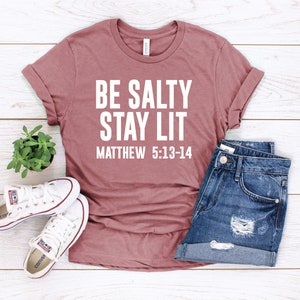 以下が含まれることがあります： 白地に「BE SALTY STAY LIT」と「MATTHEW 5:13-14」の文字が書かれたマゼンタ色のTシャツ。白い木製の表面に、白いスニーカーと青いデニムのショートパンツと一緒に置かれている。