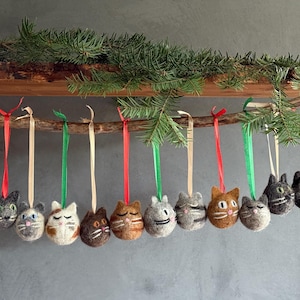 Könnte beinhalten: Handgefertigte Katzen-Weihnachtsornamente. Jedes Ornament ist eine Filzkugel mit einem Katzengesicht, aufgehängt an bunten Bändern. Die Ornamente sind an einem Holzzweig mit Tannenzweigen befestigt, ideal für die Weihnachtsdekoration.