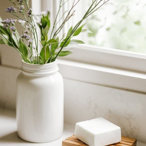 May include: A white ceramic vase filled with purple flowers sits on a white countertop next to a white bar of soap on a wooden soap dish.
