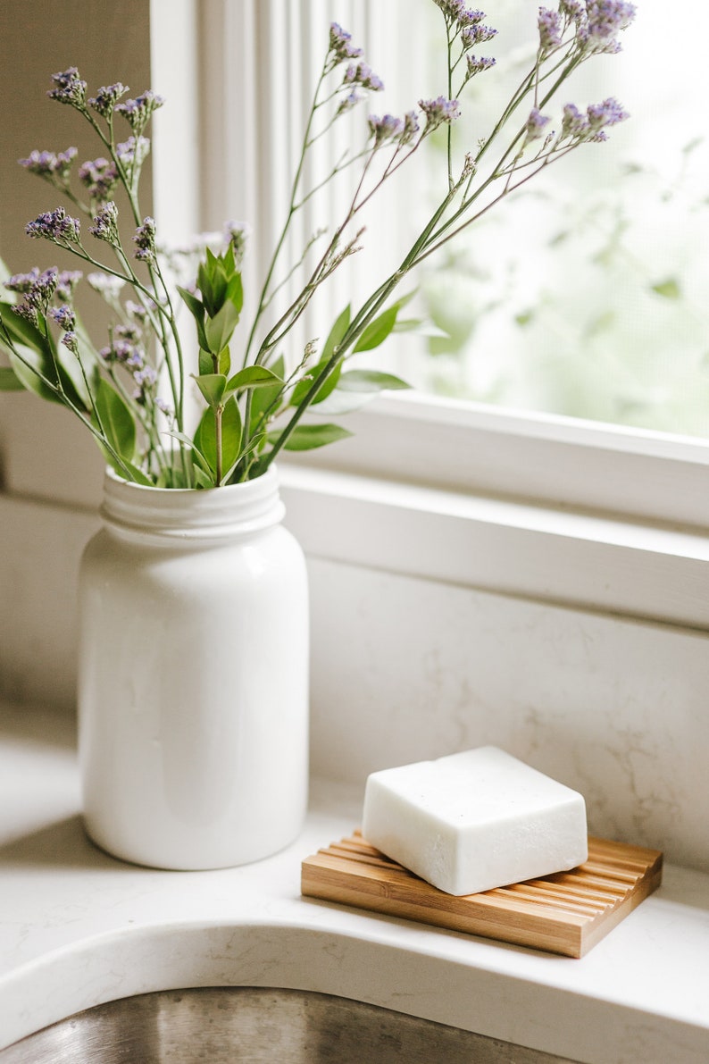 May include: A white ceramic vase filled with purple flowers sits on a white countertop next to a white bar of soap on a wooden soap dish.