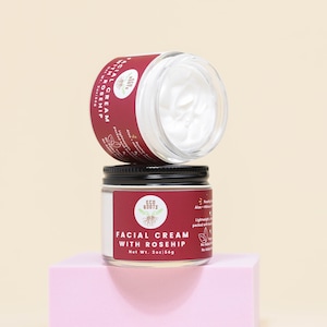 May include: Two glass jars of facial cream with black lids. The top jar is empty and the bottom jar is labeled "Facial Cream with Rosehip" and "Eco Roots". The jars are on a pink surface.