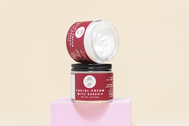 May include: Two glass jars of facial cream with black lids. The top jar is empty and the bottom jar is labeled "Facial Cream with Rosehip" and "Eco Roots". The jars are on a pink surface.