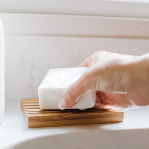 May include: A white bar of soap resting on a bamboo soap dish. The soap is slightly wet and has a few bubbles on it.