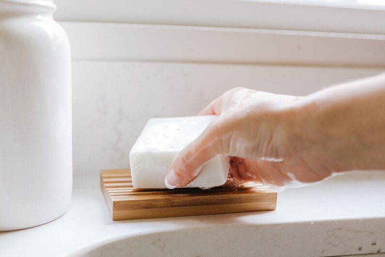 May include: A white bar of soap resting on a bamboo soap dish. The soap is slightly wet and has a few bubbles on it.