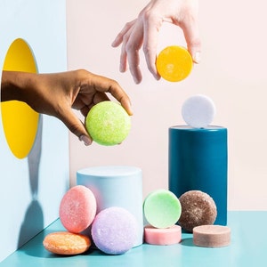 May include: A variety of colorful solid shampoo bars, including green, yellow, pink, purple, and brown, are arranged on a blue surface. Some of the bars are on a blue and white cylinder. A hand reaches into a hole in a blue wall to place a green bar on a blue cylinder.
