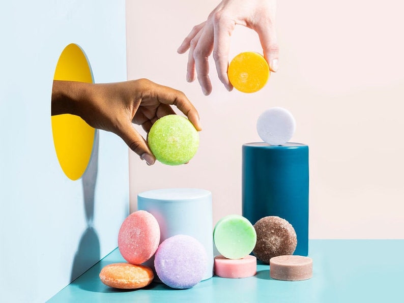 May include: A variety of colorful solid shampoo bars, including green, yellow, pink, purple, and brown, are arranged on a blue surface. Some of the bars are on a blue and white cylinder. A hand reaches into a hole in a blue wall to place a green bar on a blue cylinder.