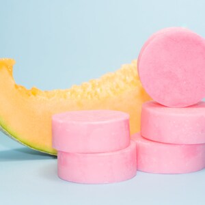 Pode incluir: Uma pilha de quatro barras de sab&atilde;o redondas cor-de-rosa sobre um fundo azul claro. Uma fatia de mel&atilde;o cantalupo est&aacute; &agrave; esquerda das barras de sab&atilde;o.