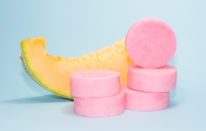 Pode incluir: Uma pilha de quatro barras de sab&atilde;o redondas cor-de-rosa sobre um fundo azul claro. Uma fatia de mel&atilde;o cantalupo est&aacute; &agrave; esquerda das barras de sab&atilde;o.