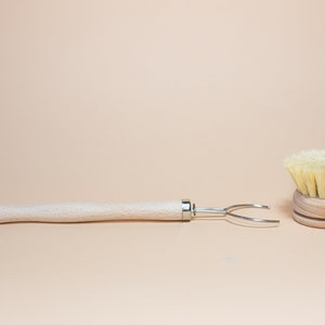 May include: A wooden dish brush with a natural bristle head and a wooden handle with a metal hook. The brush is sitting on a beige surface.