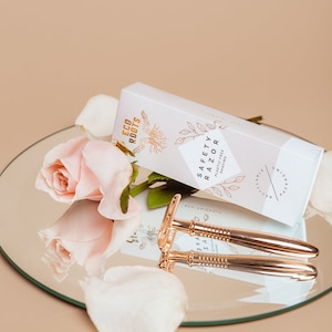 May include: A rose gold safety razor with a white box that says "Eco Roots Safety Razor Plastic Free Shaving". The razor is on a mirrored surface with a pink rose and petals.
