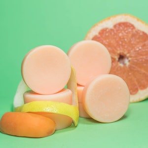 Pode incluir: Uma pilha de quatro barras de sab&atilde;o redondas de cor rosa claro com uma casca de lim&atilde;o e uma pequena fruta laranja sobre um fundo verde. Uma metade de toranja est&aacute; no fundo.