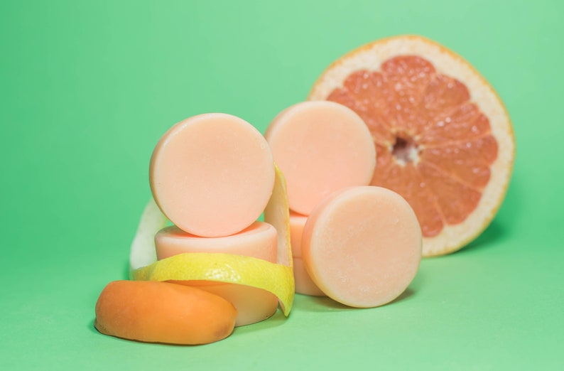Pode incluir: Uma pilha de quatro barras de sab&atilde;o redondas de cor rosa claro com uma casca de lim&atilde;o e uma pequena fruta laranja sobre um fundo verde. Uma metade de toranja est&aacute; no fundo.