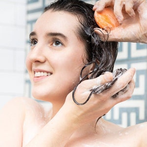 May include: A person with wet, dark hair is washing their hair with a solid orange bar of soap in the shower.