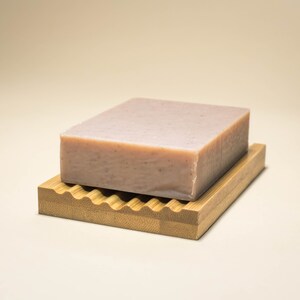 May include: A light pink bar of soap resting on a wooden soap dish with a wavy design.