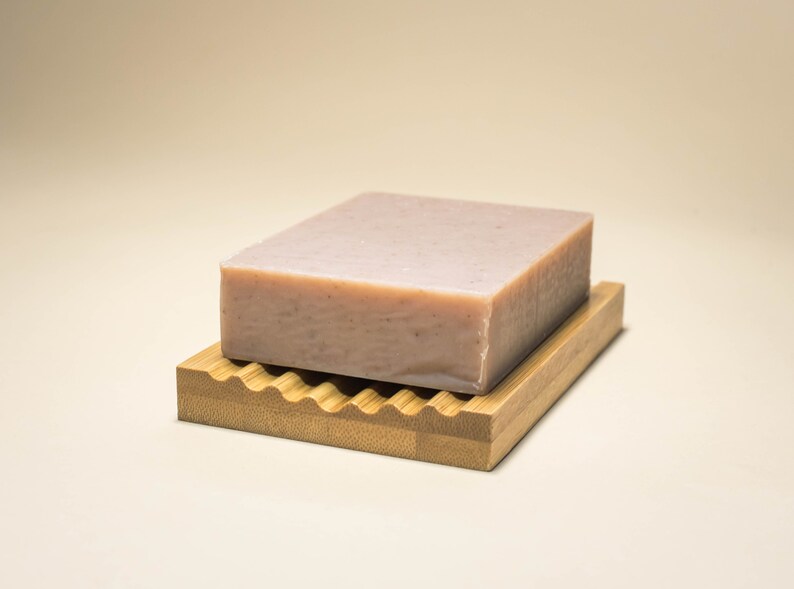 May include: A light pink bar of soap resting on a wooden soap dish with a wavy design.